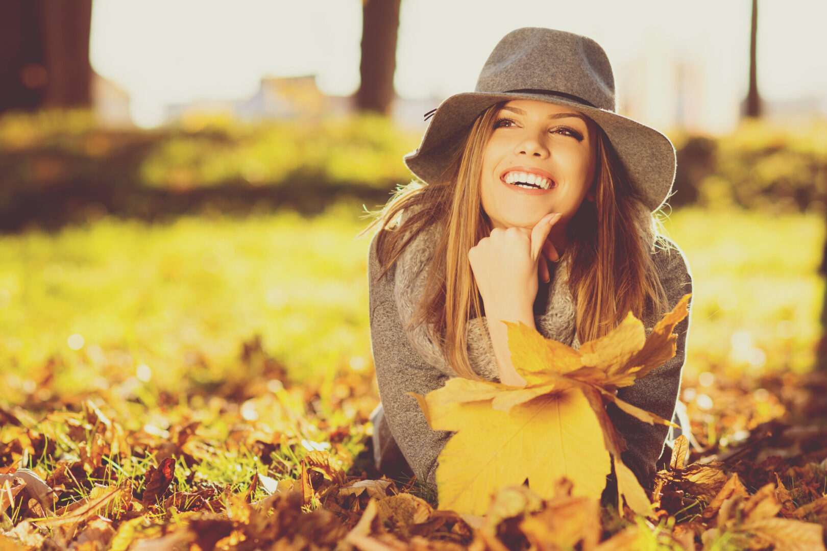 Woman in park on sunny autumn day, smiling, holding leaves.