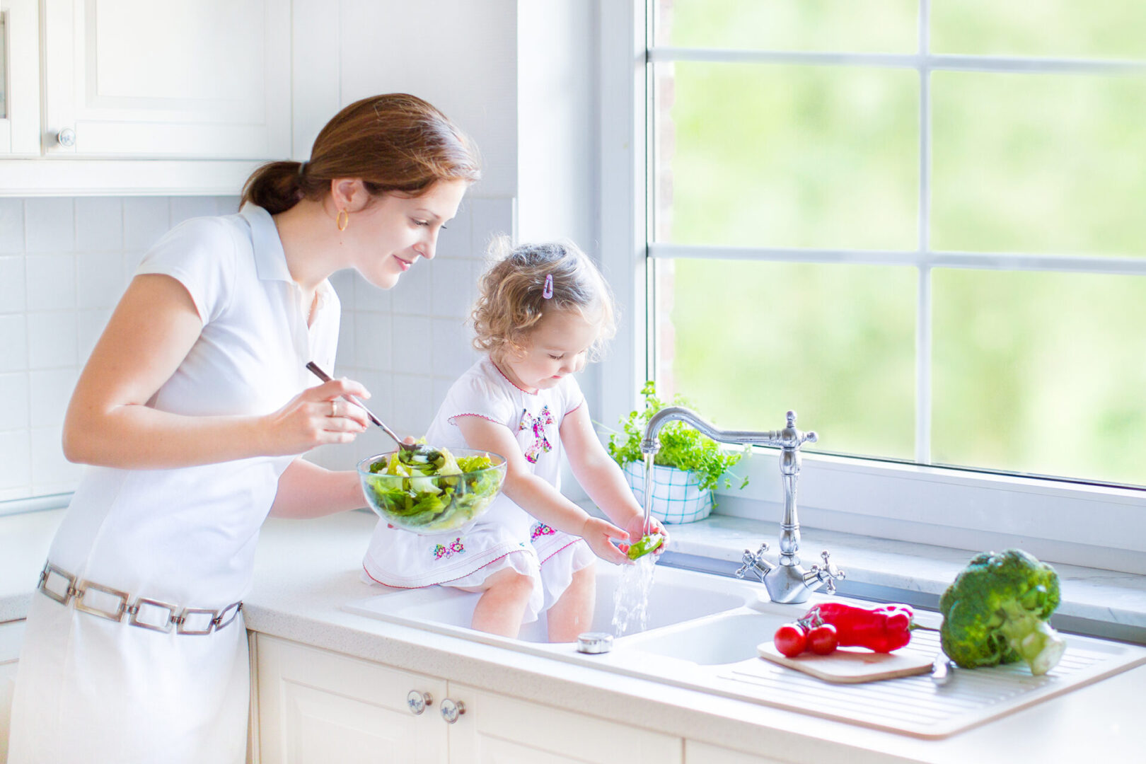 Young Beautiful Mother And Her Cute Curly-Haired Toddler Daughter Washing Vegetables Together In A Kitchen