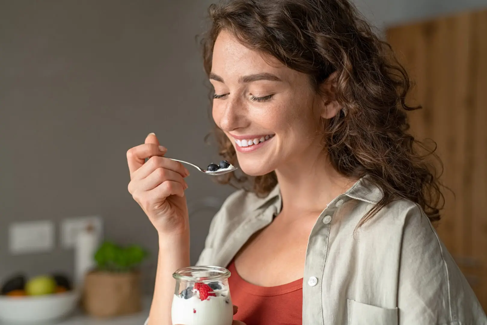 Smiling young woman having healthy breakfast at home with fruit and yogurt. Happy natural girl holding teaspoon with yogurt and blueberries. Beautiful woman eating fresh yoghurt with berries at home.