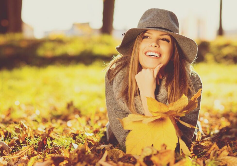 Woman in park on sunny autumn day, smiling, holding leaves.