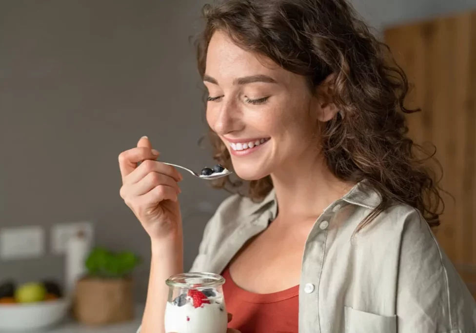 Smiling young woman having healthy breakfast at home with fruit and yogurt. Happy natural girl holding teaspoon with yogurt and blueberries. Beautiful woman eating fresh yoghurt with berries at home.